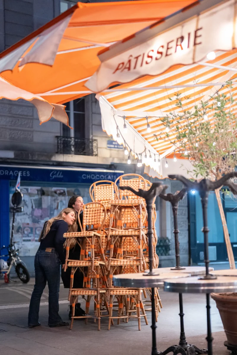 Alice · 23/04/2026 · 3896 x 5844 Cette photographie montre une scène de terrasse de pâtisserie en extérieur. Le sujet principal est une personne penchée sur un présentoir de viennoiseries en bois.La composition met en valeur les tables et chaises en bois ainsi que les parasols orange. Le cadrage est serré, centré sur la personne et le comptoir. La lumière est douce et naturelle, probablement en fin de journée. Les couleurs dominantes sont les tons chauds (orange, bois) et les bleus pastel en arrière-plan.Le style est réaliste et quotidien, évoquant une ambiance chaleureuse et conviviale, typique d’un café parisien.