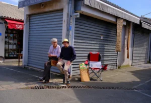 Dominique · 25/04/2026 · 7208 x 4893 Cette photographie montre deux hommes assis sur un banc devant un entrepôt aux volets métalliques fermés. Le sujet principal est leur présence détendue, avec des accessoires comme un vélo et un sac à côté.La composition est simple et symétrique, avec un cadrage serré sur les personnages et leur environnement immédiat. La lumière est naturelle, probablement en journée, avec des ombres marquées. Les couleurs dominantes sont neutres : gris des volets, beige des murs, et touches de rouge et noir sur les vêtements et objets.L’ambiance est calme et quotidienne, évoquant une pause dans une ville. Le style est réaliste et documentaire.