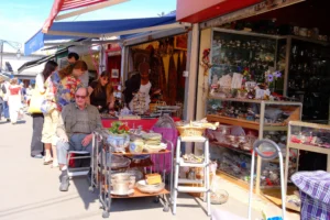 Dominique · 25/04/2026 · 6968 x 4645 Cette photographie montre un marché artisanal en extérieur. Le **sujet principal** est un stand de vente proposant des objets décoratifs et des pâtisseries.La **composition** est dynamique, avec des étals colorés et des clients en mouvement. Le **cadrage** est serré, mettant en valeur les détails des produits et les interactions humaines. La **lumière** est naturelle et claire, suggérant une journée ensoleillée.Les **couleurs dominantes** sont vives : rouges, blancs, bleus et dorés. Le **style** est réaliste et spontané, reflétant une ambiance **vivante et conviviale**, typique d’un marché local.