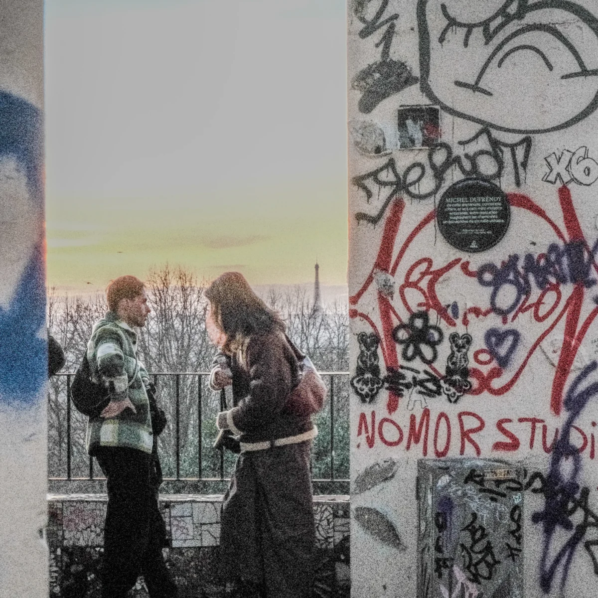 Nathalie F. · 07/02/2026 · 4160 x 4160 Cette photographie met en scène deux personnes marchant main dans la main devant un mur tagué. Le sujet principal est leur complicité et leur environnement urbain.La composition juxtapose un premier plan avec des graffitis colorés et un arrière-plan flou de ville. Le cadrage est serré sur les personnages, encadrés par les éléments architecturaux.La lumière douce, probablement crépusculaire, adoucit les contours. Les couleurs dominantes sont les tons froids (gris, bleu) et les touches chaudes des graffitis (rouge, jaune).Le style est réaliste et légèrement esthétisé, évoquant une ambiance mélancolique et poétique.