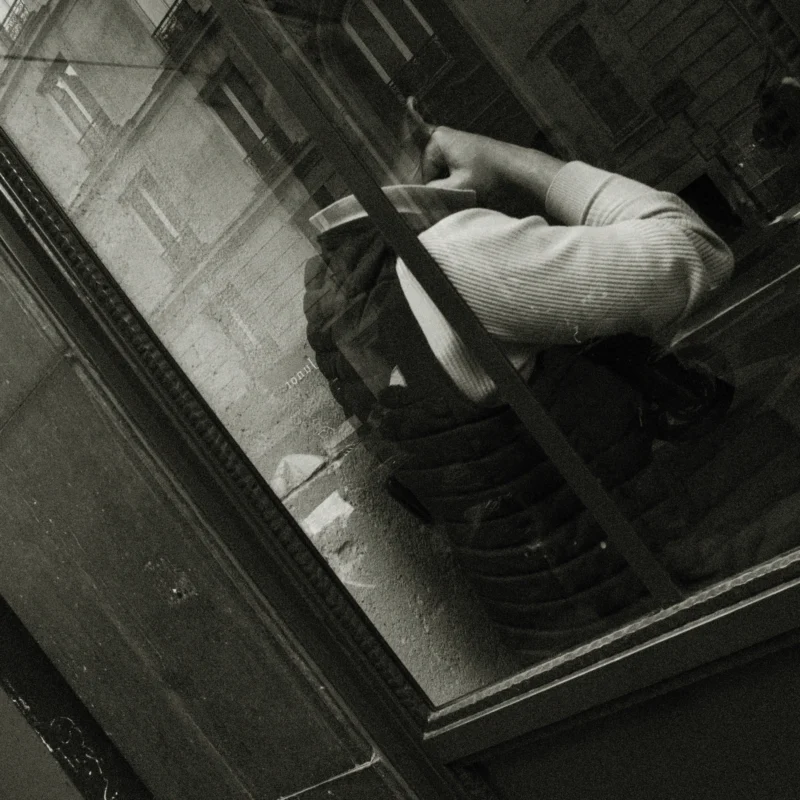 Nathalie F. · 11/04/2026 · 4160 x 4160 Cette photographie en noir et blanc montre une personne, vue de haut, penchée sur un balcon ou une fenêtre. Le sujet principal semble être cette silhouette, partiellement cachée par la structure.La composition est dynamique, avec un cadrage serré et un angle plongeant. La lumière est tamisée, créant des contrastes doux et des ombres marquées. Les couleurs dominantes, bien que limitées au noir et blanc, suggèrent des tons gris et des reflets sombres.Le style est documentaire et intime, évoquant une ambiance mélancolique et mystérieuse, renforcée par le flou et les jeux d'ombres.