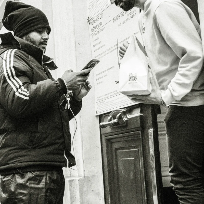 Nathalie F. · 4160 x 4160 Cette photographie en noir et blanc montre deux hommes en train de voter. Le sujet principal est l’un d’eux, concentré sur son bulletin de vote et son téléphone portable. La composition est serrée, avec un cadrage rapproché mettant en valeur leurs visages et gestes.La lumière est naturelle et semble provenir de la gauche, créant des contrastes doux. Les couleurs dominantes, bien que monochromes, jouent sur les nuances de gris et de noir, renforçant l’aspect authentique et sobre.Le style est documentaire, capturant un moment réel. L’ambiance est sérieuse et engagée, reflétant l’importance de l’acte de vote.