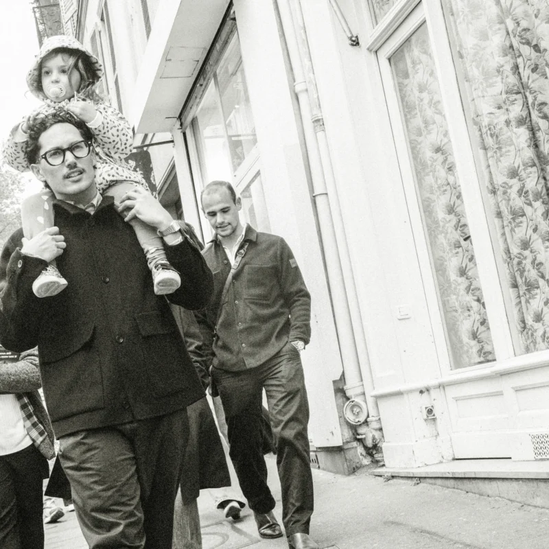 Nathalie F. · 11/04/2026 · 4160 x 4160 Cette photographie en noir et blanc capture deux hommes marchant côte à côte, l’un portant un manteau et un béret, l’autre un costume. Le sujet principal semble être ces deux individus, avec une composition centrée sur eux.Le cadrage inclut un troisième personnage en arrière-plan, partiellement visible. La lumière est naturelle, douce, et met en valeur les contours des sujets. L’ambiance est sobre et intemporelle, évoquant une scène des années 1960-1970. Le style est documentaire, avec une touche artistique. Les tons dominants sont les gris et noirs, renforçant l’aspect rétro et cinématographique.
