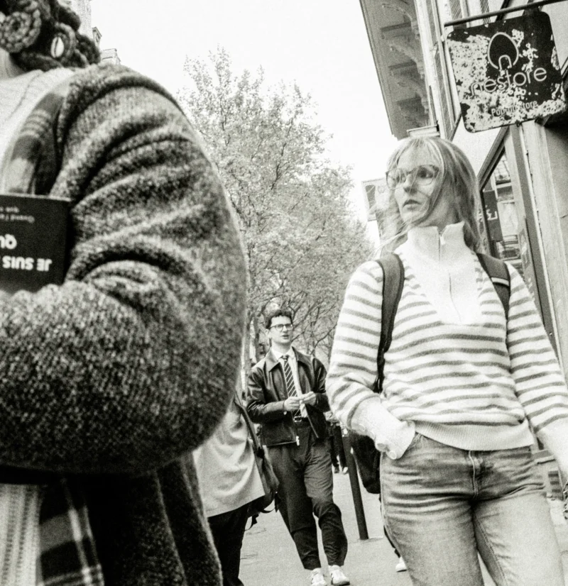 Nathalie F. · 3238 x 3343 Cette photographie en noir et blanc met en scène une femme blonde, vêtue d’un pull rayé et d’un jean, comme sujet principal. Elle semble observer quelque chose avec attention.La composition est dynamique, avec un cadrage serré sur son visage et son torse, tandis que l’arrière-plan montre des passants flous. La lumière est douce, naturelle, créant des contrastes modérés.Le style est documentaire, capturant un moment spontané. L’ambiance est urbaine et intimiste, renforcée par les tons de gris et les ombres, évoquant une scène de rue authentique et quotidienne.