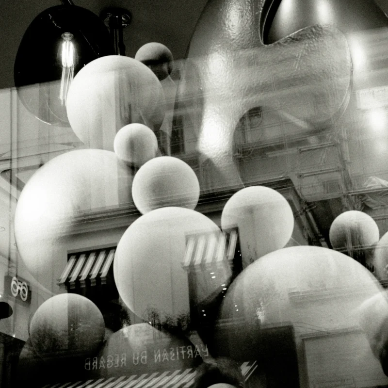 Nathalie F. · 11/04/2026 · 4160 x 4160 Cette photographie met en scène des ballons blancs flottant devant une façade architecturale, partiellement floue. Le sujet principal est l'accumulation de ces sphères, évoquant une fête ou une célébration.La composition joue sur le contraste entre les formes rondes et la structure rectiligne de l'édifice. Le cadrage serré et l'angle incliné créent une impression de profondeur et de mouvement. La lumière, douce et diffuse, accentue les textures et les ombres.Les couleurs dominantes sont les tons gris et blancs, typiques d'une image en noir et blanc. Le style est artistique et onirique, avec une ambiance à la fois nostalgique et mystérieuse.