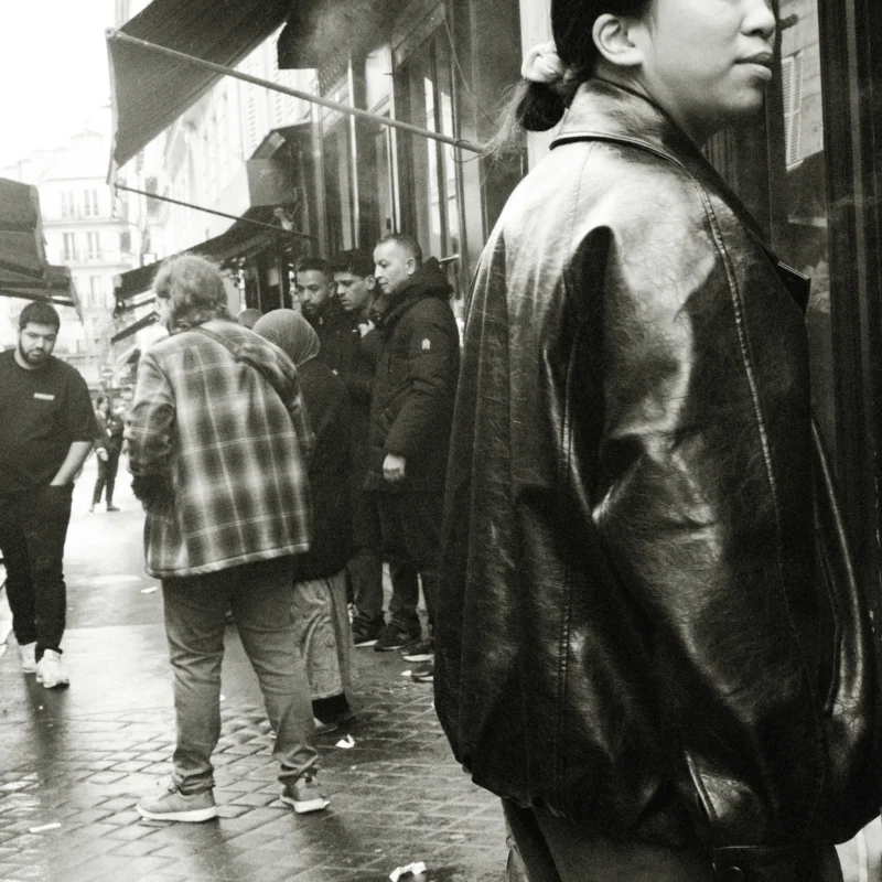 Nathalie F. · 11/04/2026 · 4160 x 4160 Cette photographie en noir et blanc met en scène un homme en manteau comme sujet principal, vu de dos, marchant dans une rue animée. La composition joue sur les contrastes entre les formes et les ombres. Le cadrage est serré sur les personnages, créant une ambiance intimiste malgré la foule.La lumière, tamisée, accentue les textures et les jeux d'ombres, renforçant le côté dramatique. Les couleurs dominantes, bien que monochromes, varient des gris clairs aux noirs profonds.Le style rappelle le photojournalisme classique, évoquant une ambiance urbaine et mélancolique, typique des scènes de vie quotidienne.