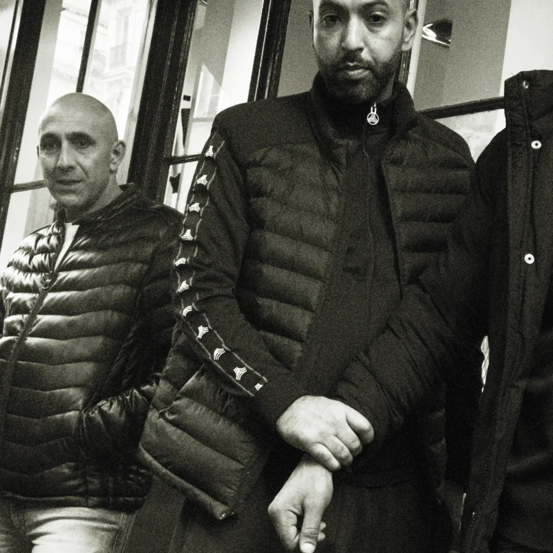 Nathalie F. · 11/04/2026 · 4160 x 4160 Cette photographie en noir et blanc met en scène deux hommes comme sujets principaux. Le cadrage est serré, centré sur eux, avec un léger flou d'arrière-plan.La composition montre un homme à gauche partiellement visible et un autre à droite, plus en détail. La lumière est douce, créant des contrastes modérés et des ombres subtiles.Les couleurs dominantes sont les nuances de gris, typiques du noir et blanc, renforçant une ambiance sobre et intemporelle. Le style est réaliste et documentaire, évoquant une ambiance urbaine et contemplative.