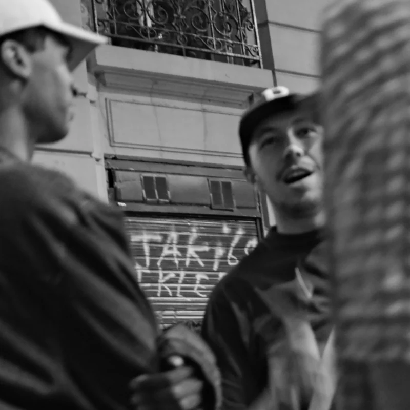 Nathalie F. · 23/04/2026 · 4160 x 4160 Cette photographie en noir et blanc met en scène deux hommes en conversation. Le sujet principal semble être l’homme au premier plan, vêtu d’une veste et d’un bonnet, regardant vers la droite. La composition est serrée, avec un cadrage rapproché qui accentue les détails des visages et des vêtements.L’ambiance est urbaine et légèrement nocturne, renforcée par les néons visibles en arrière-plan. Les couleurs dominantes sont les tons de gris, typiques du noir et blanc, créant une atmosphère à la fois moderne et intemporelle. Le style est documentaire, captant un moment authentique et spontané.