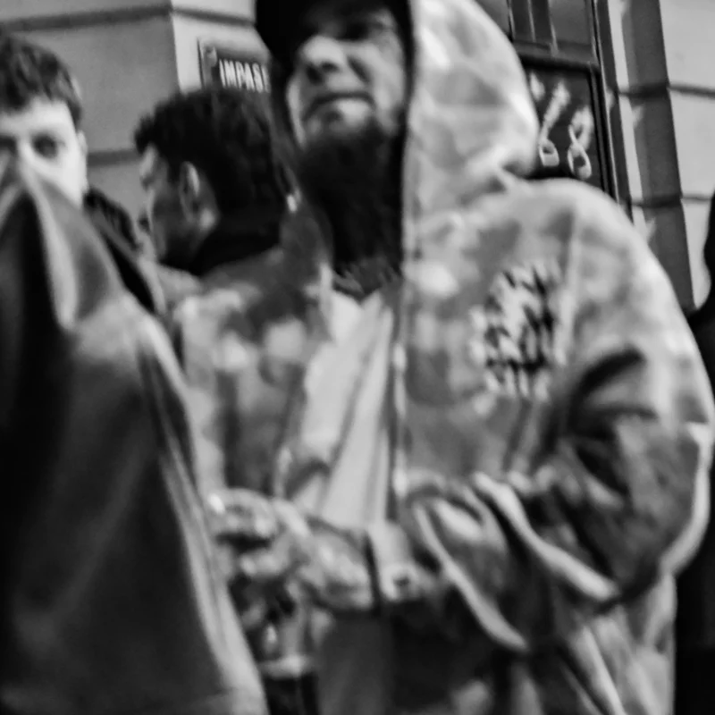 Nathalie F. · 23/04/2026 · 4160 x 4160 Cette photographie en noir et blanc met en scène un groupe de personnes dans un lieu public, probablement une gare ou un aéroport. Le sujet principal semble être un homme au centre, vêtu d’une veste camouflage, avec un regard intense et une expression marquée.La composition est serrée, avec un cadrage rapproché qui accentue les détails des visages et des vêtements. La lumière est tamisée, créant des contrastes doux et des ombres légères. L’ambiance est dynamique et légèrement tendue, renforcée par les expressions des individus. Le style est documentaire, capturant un moment spontané et authentique.