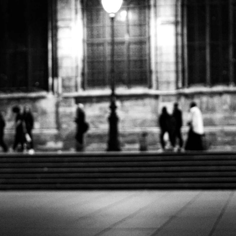 Nathalie F. · 23/04/2026 · 4160 x 4160 Cette photographie en noir et blanc capture une scène urbaine nocturne. Le sujet principal semble être un groupe de personnes en mouvement devant une façade architecturale, probablement une cathédrale ou un édifice religieux.La composition est dynamique, avec des silhouettes floues suggérant un mouvement rapide. Le cadrage est serré, centré sur les figures et les détails architecturaux. La lumière est tamisée, créant des contrastes forts et une ambiance mystérieuse. Les couleurs dominantes sont les nuances de gris, renforçant l'atmosphère nocturne et intemporelle. Le style est documentaire et artistique, évoquant une ambiance contemplative et mélancolique.
