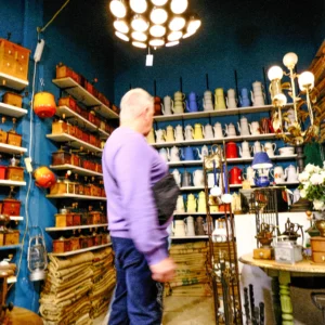 Nathalie F. · 25/04/2026 · 4160 x 4160 Cette photographie montre un homme en violet dans un magasin d’articles de pêche ou de chasse. Le sujet principal est flou, suggérant un mouvement. La composition met en valeur des étagères remplies d’objets colorés (canne à pêche, bobines, accessoires).Le cadrage est serré sur les étagères et l’homme, avec une profondeur de champ réduite. La lumière est artificielle, provenant de plusieurs sources lumineuses suspendues, créant des ombres douces. Les couleurs dominantes sont les teintes vives (rouge, bleu, vert) et les tons chauds des accessoires.L’ambiance est artisanale et chaleureuse, évoquant un magasin spécialisé et bien approvisionné.