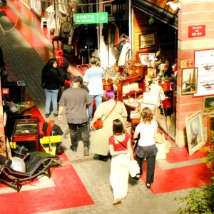 Nathalie F. · 25/04/2026 · 4160 x 4160 Cette photographie capture une scène animée d’une rue étroite et colorée. Le **sujet principal** semble être un groupe de personnes en mouvement, dont une enfant en premier plan.La **composition** est dynamique, avec des lignes convergentes vers le fond. Le **cadrage** est serré, mettant en valeur les détails des passants et des enseignes.La **lumière** est naturelle, légèrement tamisée, créant des ombres douces. Les **couleurs dominantes** sont chaudes : rouges, oranges et tons terreux, avec des éclats de blanc et noir.Le **style** est réaliste et spontané, évoquant une ambiance **vivante et populaire**, typique d’un marché ou d’une rue commerçante.