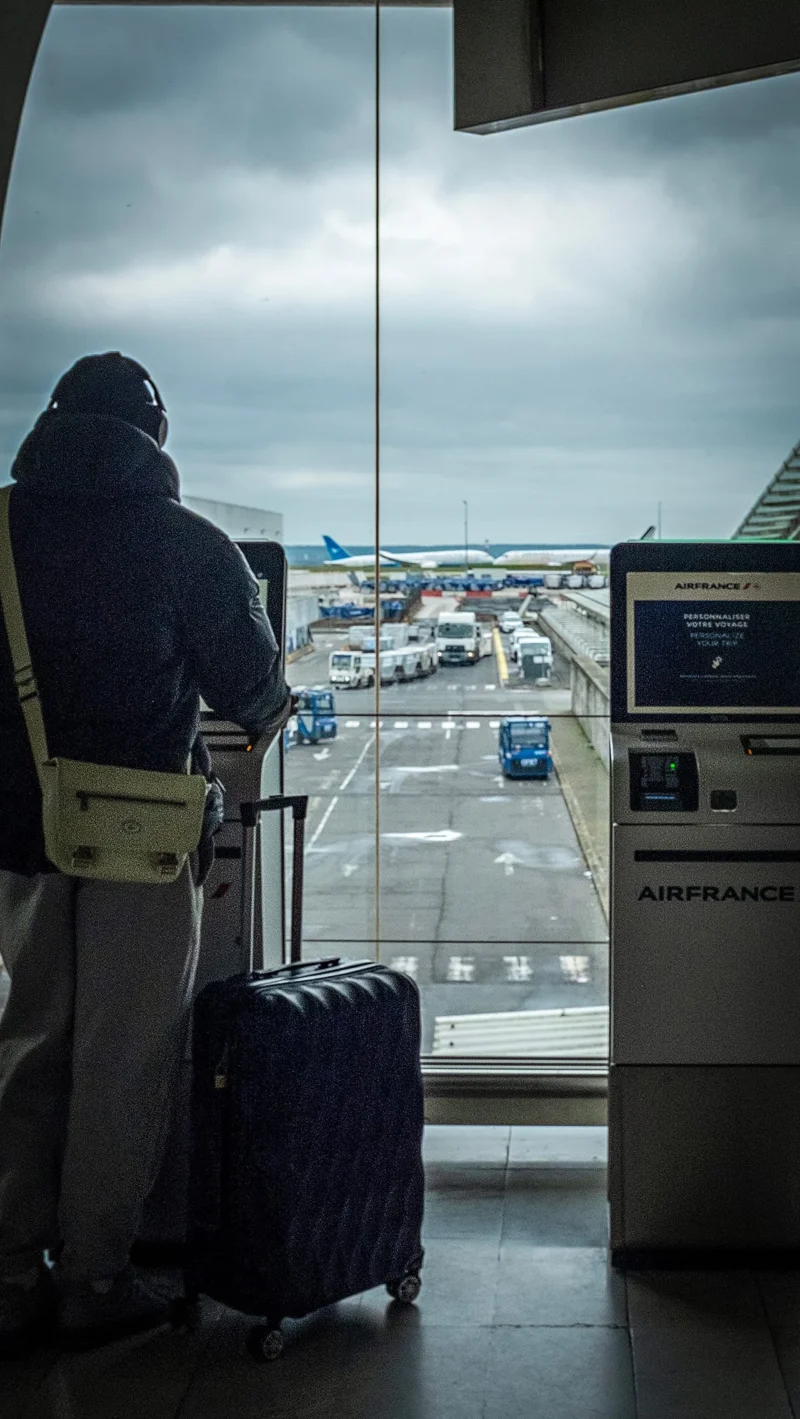 AnJo · 04/04/2026 · 1280 x 2270 Cette photographie montre un voyageur debout près d’une fenêtre d’aéroport, observant la piste. Le sujet principal est ce passager, vêtu d’un manteau sombre, avec une valise à roulettes.La composition est simple et centrée sur le contraste entre l’intérieur et l’extérieur. Le cadrage est serré sur le personnage, avec une vue dégagée sur les avions et la piste en arrière-plan.La lumière est naturelle et tamisée, créant une ambiance sobre. Les couleurs dominantes sont les gris et bleus froids, renforçant une sensation de calme et de modernité. Le style est réaliste et documentaire, évoquant une attente ou une réflexion.