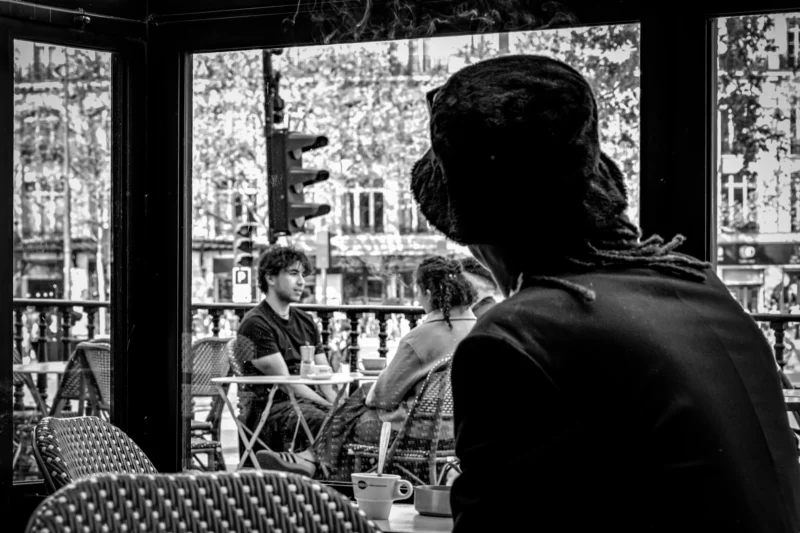 AnJo · 18/04/2026 · 5221 x 3481 Cette photographie en noir et blanc capture une scène intimiste dans un café ou un restaurant. Le sujet principal est un homme assis dos à la caméra, observant deux personnes assises à une table à l'extérieur.La composition joue sur les plans superposés, avec une profondeur de champ qui met en valeur les détails des tables et des personnes. Le cadrage est serré, centré sur les interactions entre les individus.La lumière est douce, créant des contrastes subtils et des ombres légères. Les couleurs dominantes sont les tons de gris, typiques du noir et blanc, renforçant une ambiance à la fois mélancolique et chaleureuse. Le style est documentaire et introspectif.