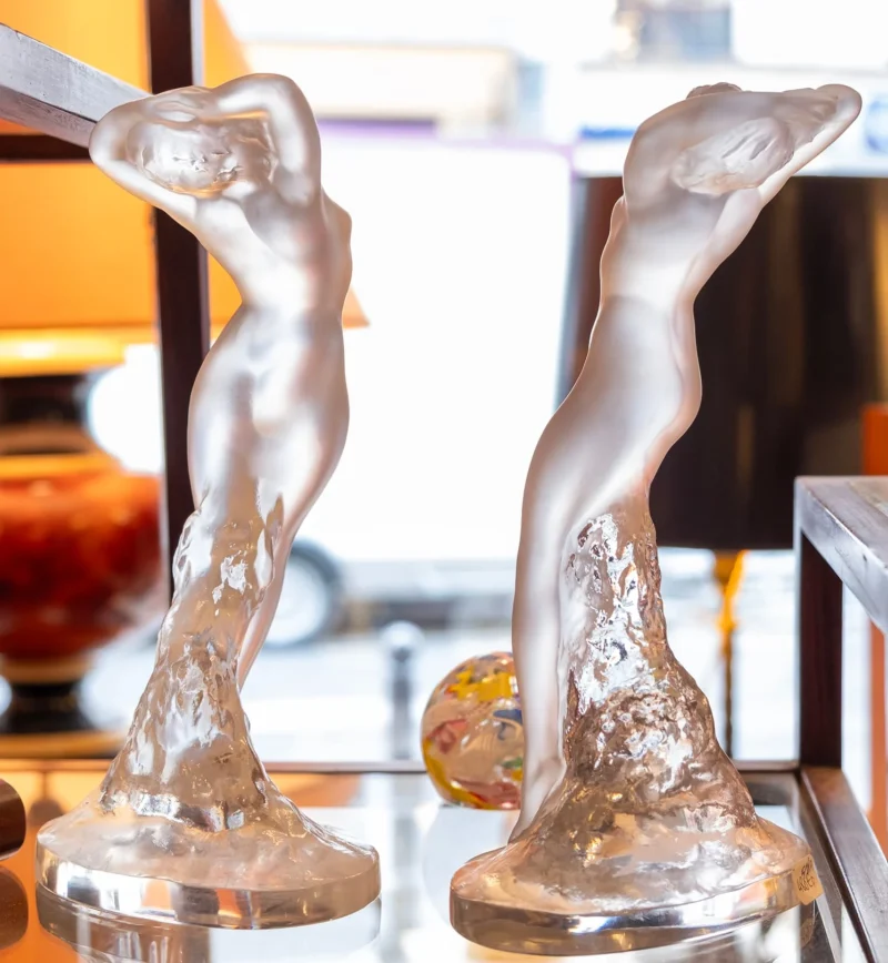 Nathalie C H · 18/04/2026 · 1493 x 1620 Cette photographie met en scène deux sculptures en cristal représentant des figures féminines stylisées, posées sur des socles. La composition est symétrique, avec un cadrage serré sur les sculptures. La lumière, douce et naturelle, provient probablement d’une source externe, éclairant les détails des formes translucides.Les couleurs dominantes sont le blanc cristallin et des teintes chaudes en arrière-plan (orange, jaune). Le style est élégant et raffiné, évoquant l’art déco ou contemporain. L’ambiance est luxueuse et sophistiquée, renforcée par les reflets et la transparence du cristal.