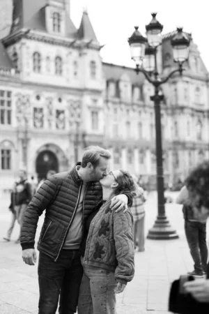Jean-Marc · 1366 x 2048 Cette photographie en noir et blanc met en scène un couple qui s'embrasse tendrement, sujet principal central. La composition est équilibrée, avec un cadrage serré sur les deux personnages, tout en intégrant un arrière-plan architectural élégant, probablement une place urbaine.La lumière est douce, naturelle, et semble provenir d'une source diffuse, créant des contrastes subtils. L'absence de couleurs accentue les textures et les jeux d'ombres.Le style est documentaire et romantique, évoquant une ambiance intime et nostalgique, renforcée par le flou léger de l'arrière-plan.