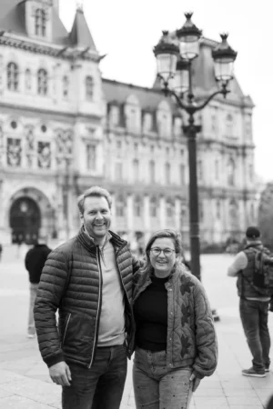 Jean-Marc · 1366 x 2048 Cette photographie en noir et blanc met en scène un couple comme sujet principal, souriant et posant côte à côte. La composition est symétrique, avec les deux personnes au premier plan, encadrées par une architecture élégante en arrière-plan, probablement un hôtel particulier ou un bâtiment historique.Le cadrage est serré sur les sujets, tout en intégrant des éléments urbains comme un lampadaire. La lumière est douce, avec des contrastes modérés, créant une ambiance chaleureuse et nostalgique. Le style est documentaire et intime, évoquant une promenade en ville. Les tons dominants, en noir et blanc, renforcent l’aspect classique et temporel de la scène.