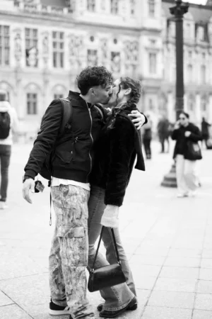 Jean-Marc · 1366 x 2048 Cette photographie en noir et blanc met en scène un couple qui s'embrasse tendrement en extérieur. Le sujet principal est au centre, avec une composition serrée qui isole les deux personnes.Le cadrage est serré sur eux, avec un arrière-plan flou montrant des bâtiments et des passants. La lumière est douce et naturelle, probablement en journée. Les tons dominants sont les gris et noirs, typiques du noir et blanc, accentuant les contrastes.Le style est documentaire et spontané, évoquant une ambiance romantique et intime dans un cadre urbain.