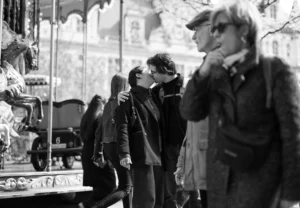 Jean-Marc · 2048 x 1417 Cette photographie en noir et blanc capture une scène de rue animée. Le sujet principal semble être un couple en train de s'embrasser, au centre de l'image. La composition montre plusieurs personnes en mouvement, créant une ambiance dynamique.Le cadrage est serré sur les personnages principaux, avec un arrière-plan légèrement flou. La lumière est douce, typique d’un jour couvert ou d’un éclairage naturel tamisé, ajoutant une touche nostalgique. Les couleurs dominantes, bien que monochromes, varient des tons clairs aux ombres profondes.Le style rappelle le cinéma classique ou documentaire, évoquant une ambiance à la fois intime et urbaine.