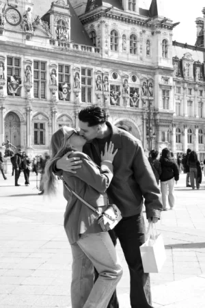 Jean-Marc · 1366 x 2048 Cette photographie en noir et blanc met en scène un couple en train de s'embrasser tendrement devant un bâtiment monumental, probablement l'Hôtel de Ville de Paris.Le **sujet principal** est le couple, au premier plan, avec une composition centrée et un **cadrage serré** qui isole leur intimité. L'arrière-plan montre des passants flous, créant un contraste entre émotion et vie urbaine.La **lumière** est naturelle, douce, et met en valeur les contours des visages. Les **couleurs dominantes** sont les nuances de gris, renforçant l'atmosphère nostalgique et élégante.Le **style** est documentaire et spontané, évoquant une **ambiance** romantique et poétique dans un cadre historique.