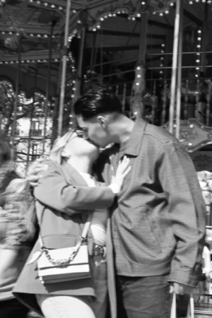 Aurélie · 15/01/2021 · 3712 x 5568 Cette photographie en noir et blanc montre deux hommes, dont l’un semble embrasser l’autre sur la joue. Le sujet principal est leur interaction intime et chaleureuse. La composition met en scène un cadre serré, centré sur eux, avec des éléments festifs en arrière-plan, comme des décorations lumineuses et des structures de manèges. La lumière est douce, créant des contrastes modérés. L’ambiance évoque une fête ou un événement joyeux, probablement dans un parc d’attractions ou une foire. Le style est documentaire, capturant un moment authentique et émouvant.
