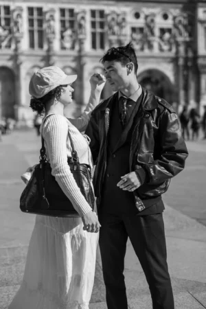 Jean-Marc · 1366 x 2048 Cette photographie en noir et blanc met en scène un couple comme sujet principal. La jeune femme, vêtue d’une robe légère et d’un chapeau, et l’homme en veste de cuir, se tiennent par la main. La composition est centrée sur eux, avec un cadrage serré qui isole leur interaction.La lumière douce et naturelle crée des contrastes subtils. L’ambiance est intime et romantique, renforcée par leur proximité et leur regard échangé. Le style évoque une touche vintage, accentuée par le traitement monochrome. L’arrière-plan flou et les bâtiments classiques ajoutent une dimension historique et élégante.
