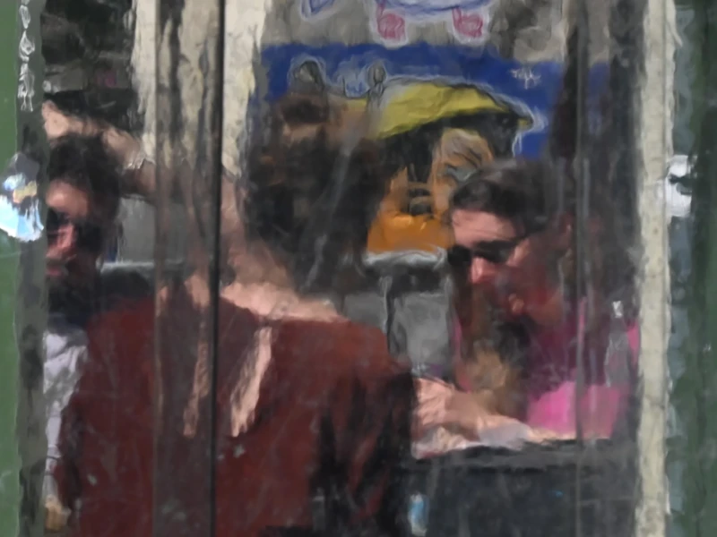 Cette photographie montre deux personnes assises dans un espace confiné, probablement un véhicule. Le sujet principal semble être leur interaction, bien que floue. La composition est serrée, avec un cadrage rapproché qui isole les personnages derrière une vitre sale.La lumière est tamisée, créant une ambiance peu nette et légèrement mélancolique. Les couleurs dominantes sont des teintes sombres et froides, avec des touches de rouge et de bleu. Le style est réaliste et spontané, évoquant une scène du quotidien. L’ambiance est intimiste et un peu mélancolique, renforcée par les reflets et la texture de la vitre.
