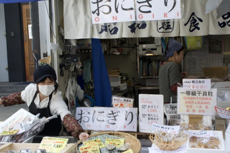 Dominique · 11/04/2024 · 6000 x 4000 Cette photographie montre un stand de rue vendant des produits alimentaires japonais, notamment des *onigiri* (boulettes de riz). Le sujet principal est la vendeuse assise, entourée de ses marchandises.La composition est serrée, mettant en avant les produits et les panneaux en japonais. Le cadrage est horizontal, captant l’ambiance d’un marché traditionnel. La lumière est naturelle et tamisée, créant une ambiance chaleureuse et authentique. Les couleurs dominantes sont neutres (beiges, bruns) avec des touches de bleu et de blanc sur les enseignes. Le style est documentaire, reflétant une scène de vie quotidienne. L’ambiance est conviviale et artisanale.