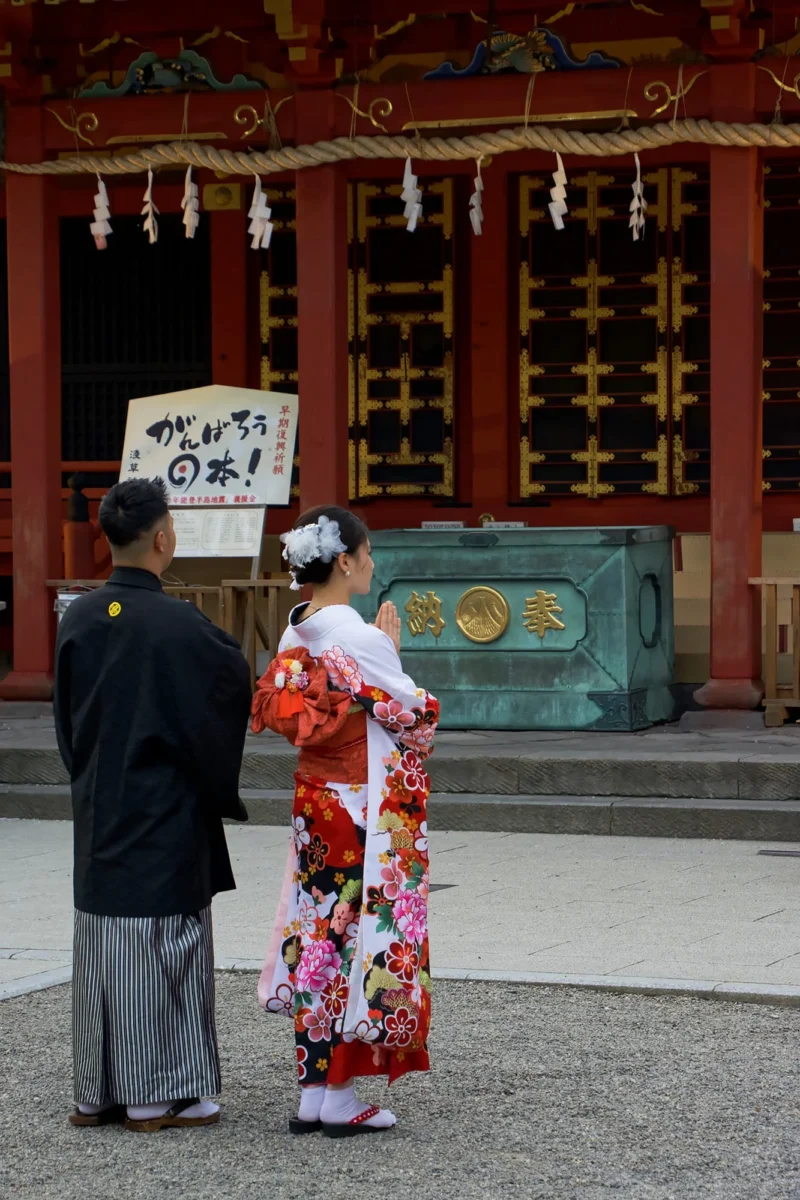 Dominique · 11/04/2024 · 1333 x 2000 Cette photographie met en scène deux personnes devant un temple japonais. Le sujet principal est une femme vêtue d’un kimono traditionnel coloré (rose et blanc), accompagnée d’un homme en tenue sombre. La composition montre une architecture typique avec des lanternes et des portes en bois rouge.Le cadrage est serré sur les personnages, avec un arrière-plan flou mais identifiable. La lumière est naturelle, douce et légèrement tamisée, créant une ambiance calme. Les couleurs dominantes sont les tons chauds (rouge, rose) et les contrastes entre les vêtements traditionnels et le bois sombre. Le style évoque la culture japonaise, avec une ambiance paisible et respectueuse.