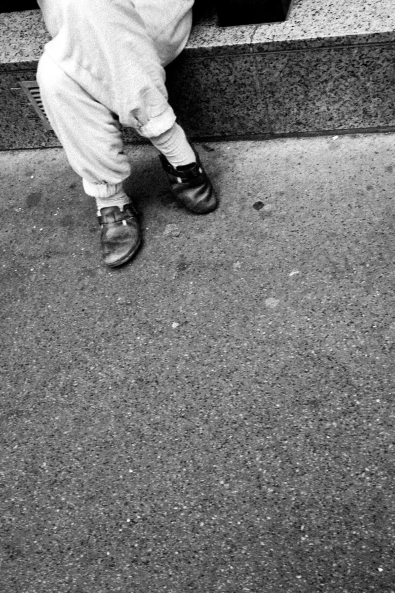Daniel · 11/04/2026 · 3205 x 4808 Cette photographie en noir et blanc met en scène les pieds et les bas d’une personne vêtue de pantalons blancs et de chaussures noires. Le cadrage est serré, centré sur les pieds posés sur une surface pavée.La composition est épurée, avec un jeu d’ombres et de lumière naturelle, créant des contrastes marqués. L’ambiance est sobre et minimaliste, évoquant une atmosphère urbaine et intime. Le style photographique rappelle un documentaire social, avec une touche de réalisme brut. Les tons dominants sont les gris et noirs, renforçant l’aspect monochrome et dramatique.