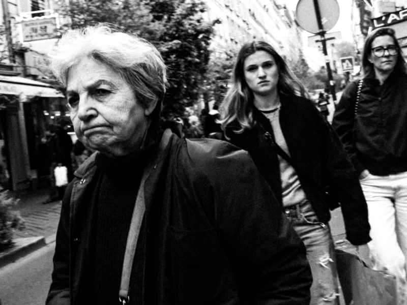 Cette photographie en noir et blanc met en scène un homme âgé comme sujet principal, marchant avec un regard pensif. La composition montre aussi deux jeunes femmes en arrière-plan, légèrement floues.Le cadrage est serré sur l'homme, avec une profondeur de champ qui isole son visage. La lumière est naturelle, douce mais contrastée, créant des ombres marquées. Les tons dominants sont les noirs, gris et blancs, typiques du noir et blanc.Le style est documentaire et réaliste, évoquant une ambiance urbaine et introspective, presque mélancolique.