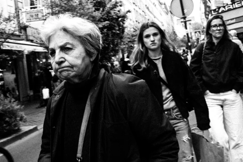 Daniel · 11/04/2026 · 3517 x 2345 Cette photographie en noir et blanc met en scène un homme âgé comme sujet principal, marchant avec un regard pensif. La composition montre aussi deux jeunes femmes en arrière-plan, légèrement floues.Le cadrage est serré sur l'homme, avec une profondeur de champ qui isole son visage. La lumière est naturelle, douce mais contrastée, créant des ombres marquées. Les tons dominants sont les noirs, gris et blancs, typiques du noir et blanc.Le style est documentaire et réaliste, évoquant une ambiance urbaine et introspective, presque mélancolique.