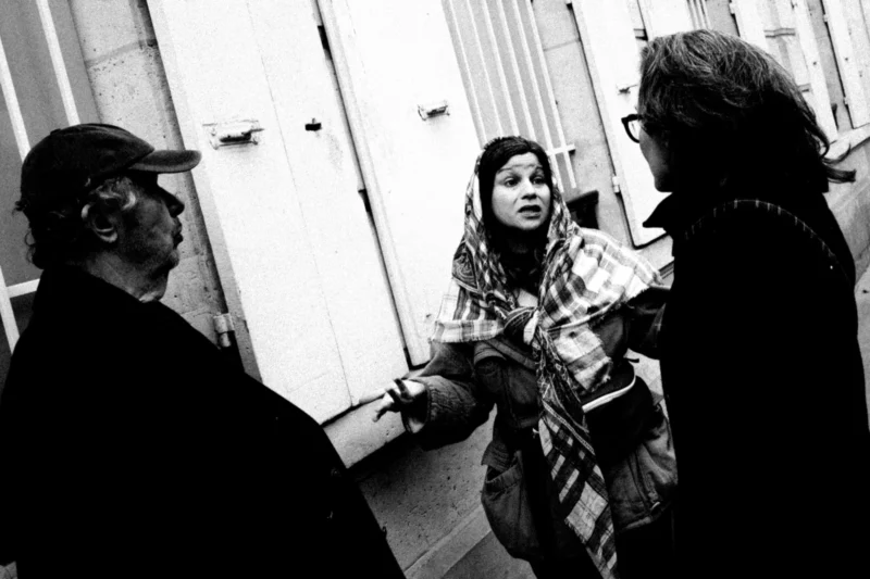 Daniel · 11/04/2026 · 4007 x 2671 Cette photographie en noir et blanc montre trois personnes en conversation dans un cadre urbain. Le sujet principal semble être la femme au centre, vêtue d’un pull à carreaux, entourée de deux hommes.La composition est serrée, avec un cadrage rapproché qui met l’accent sur les interactions. La lumière est naturelle, contrastée, créant des ombres marquées. Les tons dominants sont les gris et noirs, typiques du monochrome.Le style est documentaire, spontané, évoquant une ambiance authentique et intime, presque théâtrale. L’atmosphère suggère une discussion animée dans un environnement urbain.