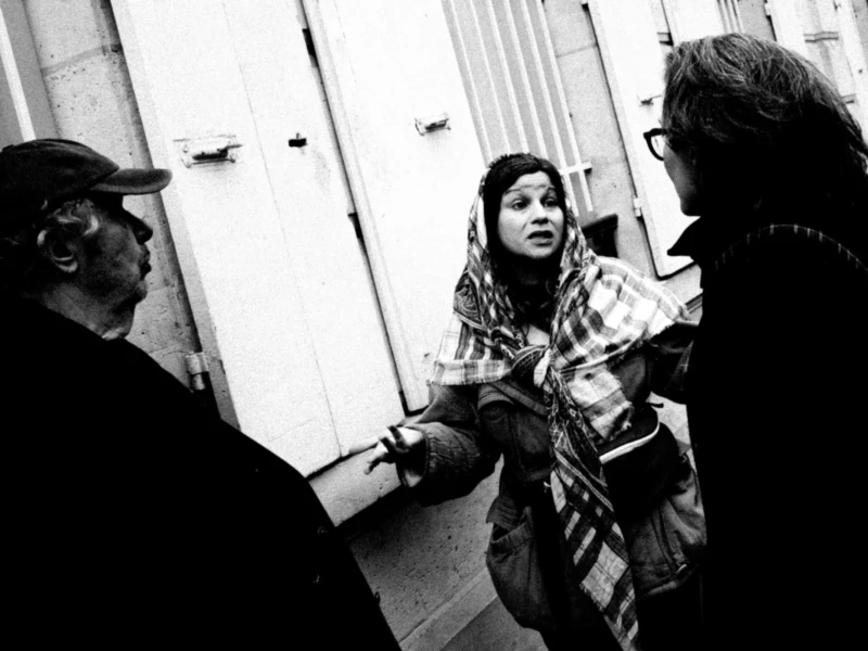 Daniel · 11/04/2026 · 4007 x 2671 Cette photographie en noir et blanc montre trois personnes en conversation dans un cadre urbain. Le sujet principal semble être la femme au centre, vêtue d’un pull à carreaux, entourée de deux hommes.La composition est serrée, avec un cadrage rapproché qui met l’accent sur les interactions. La lumière est naturelle, contrastée, créant des ombres marquées. Les tons dominants sont les gris et noirs, typiques du monochrome.Le style est documentaire, spontané, évoquant une ambiance authentique et intime, presque théâtrale. L’atmosphère suggère une discussion animée dans un environnement urbain.
