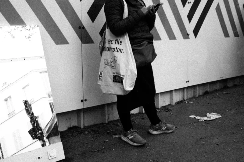 Daniel · 11/04/2026 · 4032 x 2688 Cette photographie en noir et blanc met en scène une personne de dos, tenant un sac à provisions. Le sujet principal est flou, attirant l’attention sur le sac blanc avec des motifs graphiques.La composition est simple, avec un cadrage serré sur les jambes et le bas du corps. La lumière semble naturelle, douce et diffuse, créant des contrastes modérés. Les couleurs dominantes, bien que monochromes, jouent sur les nuances de gris et de blanc.Le style est documentaire et urbain, évoquant une ambiance quotidienne et discrète, presque intimiste malgré l’absence de détails faciaux.