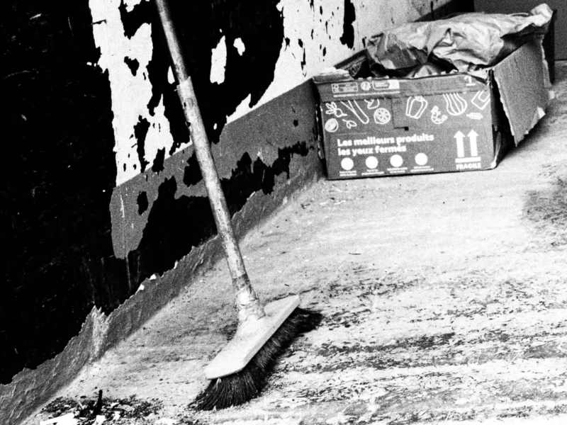 Daniel · 11/04/2026 · 2233 x 1675 Cette photographie en noir et blanc montre un environnement industriel ou artisanal. Le sujet principal est un balai et une boîte de produits chimiques (marque "NYO") posée sur une surface abîmée et sale.La composition met en avant ces éléments avec un cadrage serré, accentuant le côté utilitaire et usé. La lumière semble naturelle mais contrastée, créant des ombres marquées. Les couleurs dominantes sont les tons de gris, renforçant l’aspect vieilli et industriel. Le style est documentaire, évoquant une ambiance austère et réaliste, presque mélancolique.