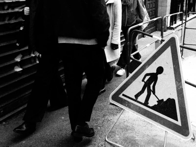 Daniel · 11/04/2026 · 3611 x 2708 Cette photographie en noir et blanc montre un **sujet principal** flou, une personne marchant près d’un panneau de signalisation triangulaire représentant un travailleur avec un marteau, indiquant des **chantiers**.La **composition** est simple, avec un cadrage serré sur les pieds et le panneau. La **lumière** est naturelle, contrastée, créant des ombres marquées. Les **couleurs dominantes** sont les nuances de gris, renforçant l’aspect urbain et industriel.Le **style** est documentaire et réaliste, avec une **ambiance** sobre et moderne, évoquant la vie quotidienne en ville.