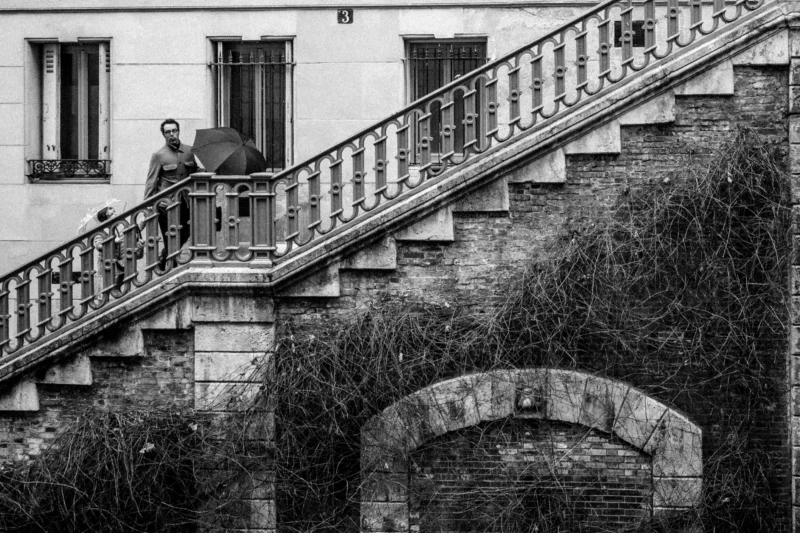 Daniel · 11/04/2026 · 4376 x 2917 Cette photographie en noir et blanc met en scène un homme assis sur une balustrade en pierre, au sommet d’un escalier extérieur. La composition joue sur la perspective ascendante, renforcée par les marches et les balustres en fer forgé. Le cadrage inclut une partie de la façade d’un bâtiment et un arbre à droite, encadrant le sujet.La lumière douce et contrastée accentue les textures et les ombres, créant une ambiance mélancolique et introspective. Les tons dominants, du gris au noir, soulignent le style artistique et dramatique, évoquant une atmosphère à la fois calme et légèrement nostalgique.