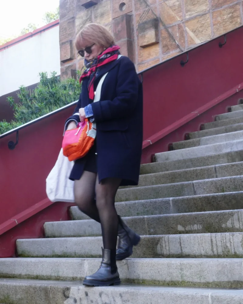 Bao · 2547 x 3186 Cette photographie montre une femme comme sujet principal, descendant un escalier extérieur. Elle porte un manteau noir, un collant résille et des chaussures plates, tenant un sac orange et un objet blanc.La composition est dynamique, avec un cadrage serré sur les jambes et le bas du corps. L’escalier rouge et les murs en pierre encadrent la scène. La lumière semble naturelle, un peu diffuse, créant des ombres douces. Les couleurs dominantes sont les tons froids (noir, gris) contrastant avec le rouge vif de l’escalier et l’orange du sac.L’ambiance est urbaine et légèrement mélancolique.