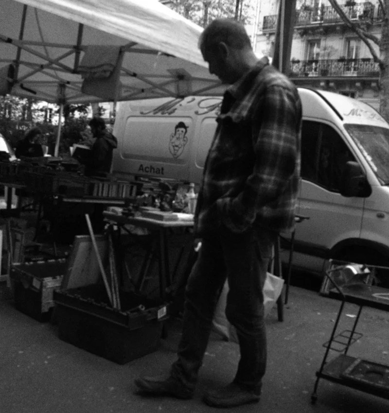 Bao · 3641 x 3866 Cette photographie en noir et blanc met en scène un homme comme sujet principal, marchant près d’un van décoré. La composition est dynamique, avec des éléments urbains en arrière-plan : étals, personnes et objets divers.Le cadrage est serré sur l’homme, mais inclut aussi des détails du marché. La lumière semble naturelle, avec des contrastes marqués créant des ombres nettes. L’ambiance est réaliste et quotidienne, évoquant une scène de rue animée. Le style est documentaire, capturant un instant authentique dans un environnement urbain.