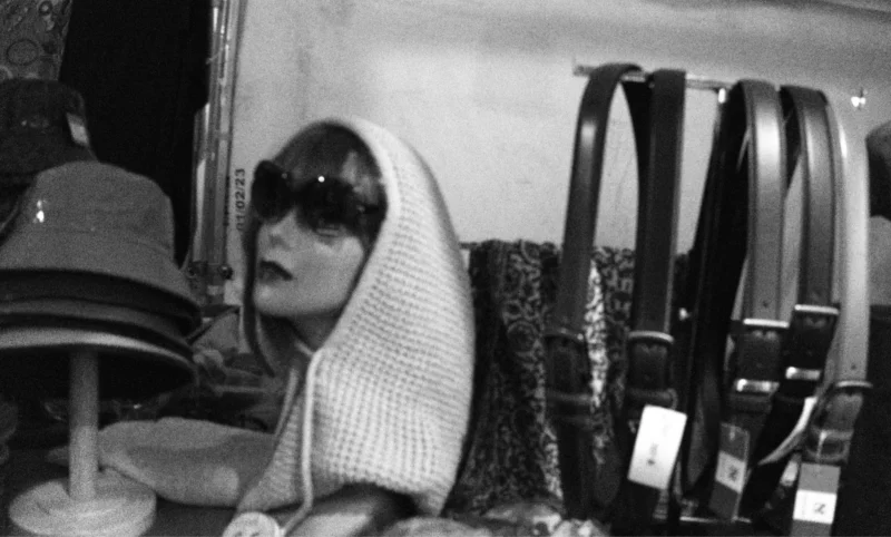 Bao · 5572 x 3355 Cette photographie en noir et blanc montre une personne, sujet principal, vêtue d’un pull et d’un foulard, les yeux partiellement cachés par des lunettes et une coiffure couvrant son visage. La composition met en avant un intérieur minimaliste, avec des étagères remplies de ceintures en arrière-plan.Le cadrage est serré, centré sur le visage et la partie supérieure du corps. La lumière semble douce et tamisée, créant des contrastes modérés. L’ambiance est intime et légèrement mystérieuse, renforcée par l’absence de couleurs et les ombres discrètes. Le style évoque un réalisme poétique, typique des clichés d’inspiration documentaire ou artistique des années 1970-1980.