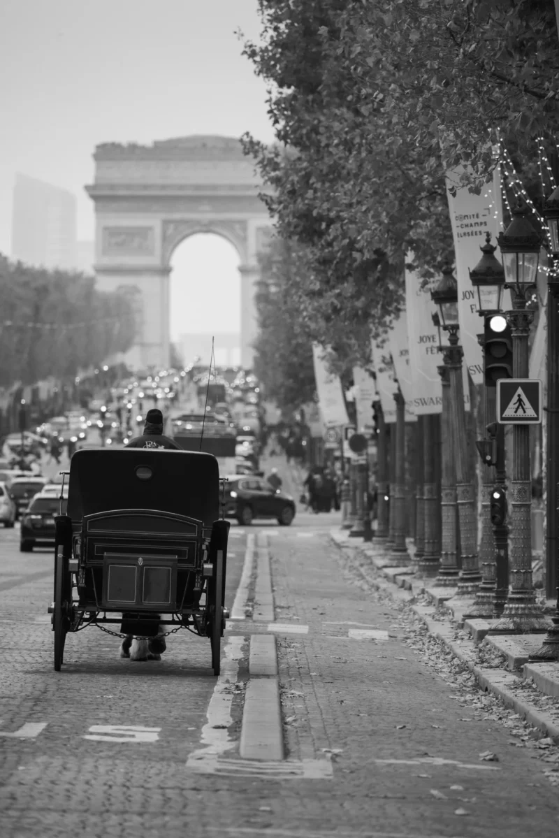 Philippe · 22/11/2025 · 2001 x 3000 Cette photographie en noir et blanc capture l'Arc de Triomphe à Paris. Le sujet principal est une voiture à cheval circulant sur les Champs-Élysées. La composition montre une perspective large, avec des arbres alignés et une foule en arrière-plan. Le cadrage est dynamique, mettant en valeur la profondeur de la scène.La lumière est douce, typique d’un jour nuageux, créant des contrastes modérés. Les couleurs dominantes sont les nuances de gris, renforçant l’aspect historique et nostalgique. Le style est documentaire, évoquant une ambiance rétro et pittoresque, mêlant tradition et vie urbaine.