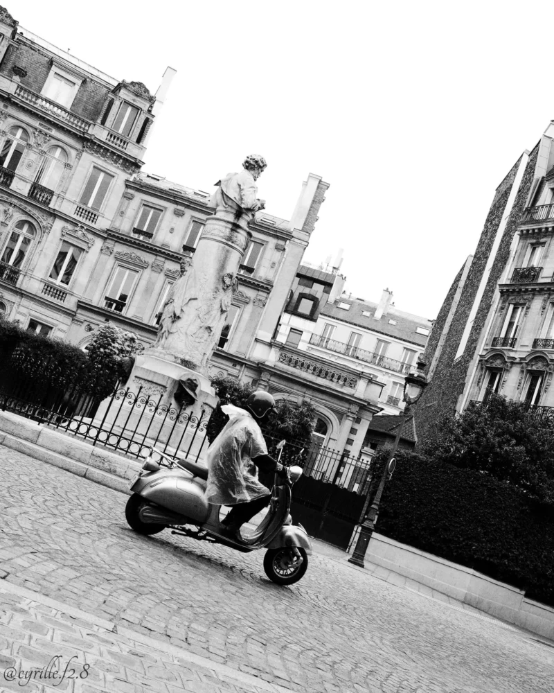 Cyrille · 11/04/2026 · 1621 x 2026 Cette photographie en noir et blanc met en scène un homme assis sur un scooter comme sujet principal. La composition montre une rue urbaine avec des bâtiments classiques en arrière-plan et une statue visible à gauche.Le cadrage est serré sur le scooter et son conducteur, tout en intégrant l’environnement architectural. La lumière est douce, sans ombres marquées, créant une ambiance sobre et légèrement nostalgique. Les tons dominants sont les gris et noirs, typiques du noir et blanc, renforçant l’aspect intemporel de la scène. Le style est documentaire et réaliste, évoquant une atmosphère calme et urbaine.