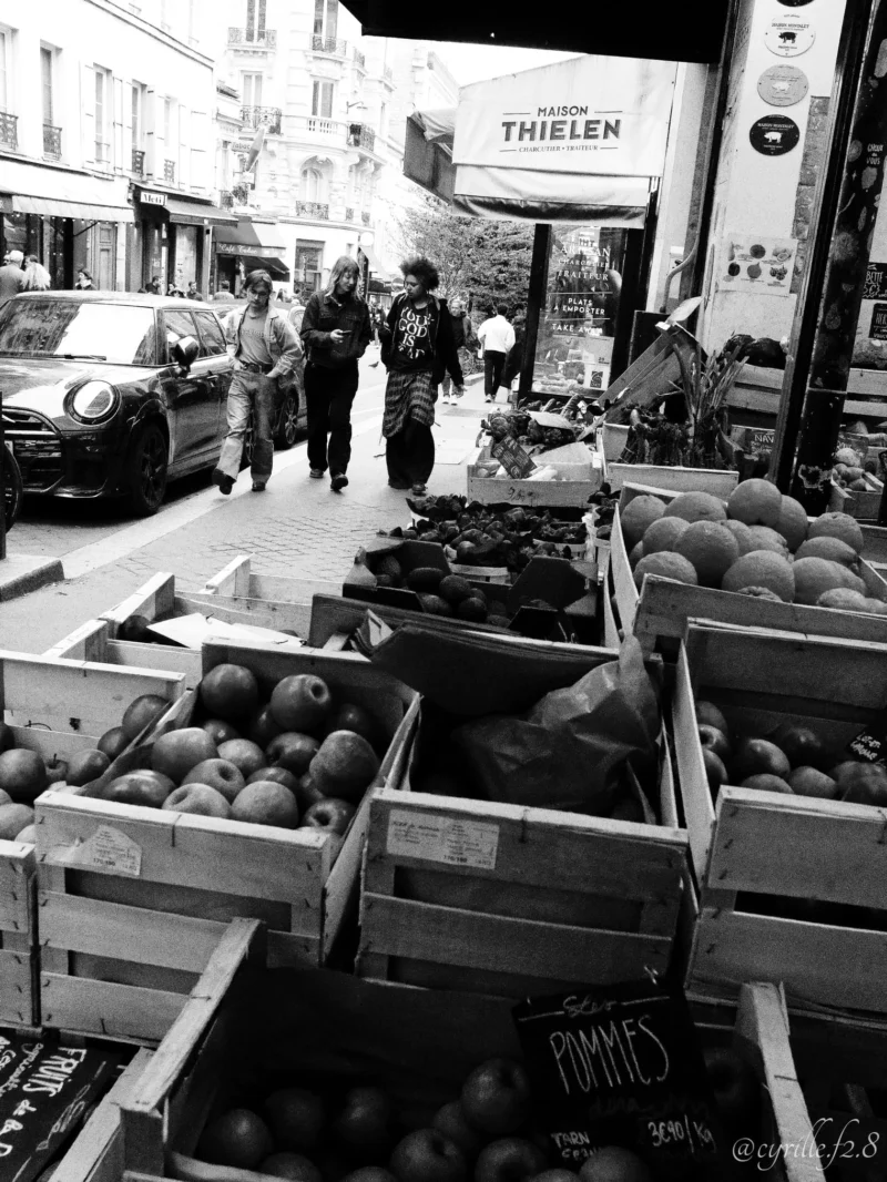 Cyrille · 11/04/2026 · 1520 x 2026 Cette photographie en noir et blanc capture un marché de fruits, principalement des melons. Le sujet principal est les étals de produits frais, avec des caisses en bois remplies de melons.La composition montre une rue animée avec des passants et des voitures en arrière-plan. Le cadrage est large, incluant une partie de la rue et des bâtiments. La lumière semble naturelle, avec des contrastes marqués créant des ombres nettes.L’ambiance est authentique et vivante, évoquant une scène quotidienne dans un marché local. Le style est documentaire et réaliste, renforcé par le traitement en noir et blanc.