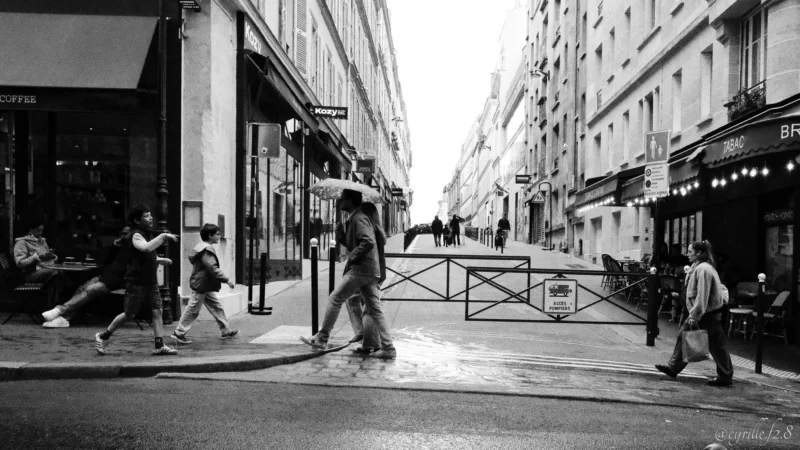Cyrille · 11/04/2026 · 2026 x 1140 Cette photographie en noir et blanc capture une scène urbaine animée. Le sujet principal semble être la rue et ses passants, dont une personne traversant avec un parapluie ouvert. La composition montre une perspective légèrement en contre-plongée, avec des bâtiments bordant la rue et des commerces aux enseignes visibles.Le cadrage est serré, mettant en valeur les détails des passants et de l’architecture. La lumière est douce, typique d’un jour couvert, créant des contrastes modérés. L’ambiance est dynamique et quotidienne, reflétant une ville active malgré la pluie. Le style est documentaire, réaliste et dépouillé.
