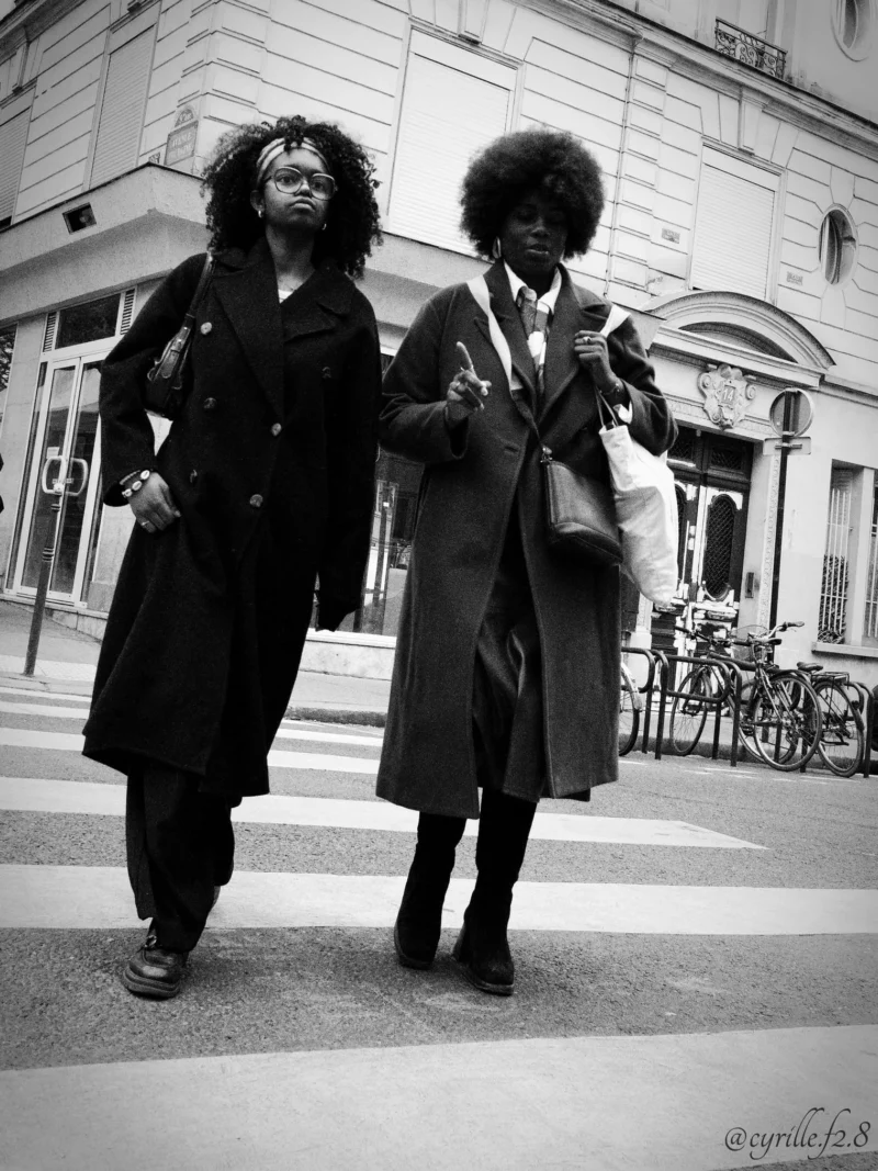 Cyrille · 11/04/2026 · 1519 x 2026 Cette photographie en noir et blanc met en scène deux personnes comme sujets principaux, marchant côte à côte. Leur style vestimentaire rétro, avec des manteaux longs et des coiffures afro, est marqué.La composition est symétrique et dynamique, avec un cadrage serré sur les sujets. L’ambiance est urbaine, suggérée par l’architecture en arrière-plan et les bancs. La lumière semble naturelle, douce et légèrement contrastée, accentuant les contours.Les tons dominants sont les gris et noirs, renforçant un style vintage et intemporel. L’ambiance générale est à la fois élégante et décontractée, évoquant une promenade dans un quartier historique.