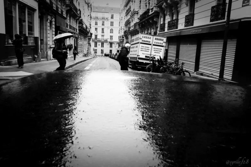 Cyrille · 11/04/2026 · 2026 x 1347 Cette photographie en noir et blanc capture une rue étroite et humide sous la pluie. Le sujet principal est une personne tenant un parapluie, marchant vers l’arrière-plan. La composition met en valeur la perspective avec des bâtiments de chaque côté convergeant vers l’horizon.Le cadrage est serré, accentuant l’effet de profondeur. La lumière est tamisée, créant des contrastes doux et des reflets sur le sol mouillé. Les couleurs dominantes, bien que monochromes, jouent sur les nuances de gris et de noir, renforçant l’ambiance mélancolique et calme. Le style est documentaire et artistique, évoquant une atmosphère contemplative et légèrement nostalgique.