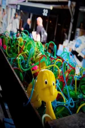 Cyrille · 25/04/2026 · 4000 x 6016 Cette photographie met en scène un éléphant en plastique jaune, sujet principal, entouré de multiples élastiques colorés emmêlés. La composition est dynamique, avec un cadrage serré sur l'éléphant, créant un effet de désordre joyeux. La lumière semble naturelle, bien que légèrement tamisée, mettant en valeur les couleurs vives des élastiques (verts, bleus, rouges) et l'objet jaune. Les tons dominants sont éclatants et contrastés. Le style est spontané et ludique, évoquant une ambiance festive et créative, presque artistique. L'arrière-plan flou suggère une activité ou un marché.
