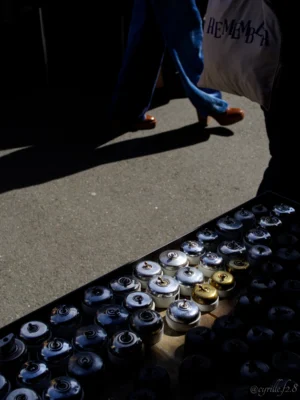 Cyrille · 25/04/2026 · 3564 x 4752 Cette photographie montre une personne marchant près d’un mémorial improvisé. Le sujet principal est l’ombre de cette personne, contrastant avec une série de bougies et de canettes posées au sol, évoquant un hommage ou une commémoration.La composition utilise un cadrage bas, mettant en valeur les objets au premier plan. La lumière naturelle crée des ombres marquées, renforçant le côté dramatique. Les couleurs dominantes sont les tons froids du béton et les reflets chauds des bougies.Le style est réaliste et documentaire, avec une ambiance mélancolique et solennelle.