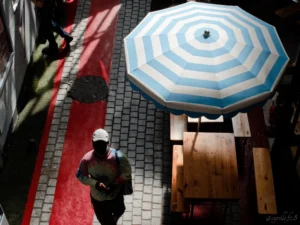 Cyrille · 25/04/2026 · 4633 x 3475 Cette photographie présente un sujet principal : un homme en tenue de travail (casque blanc) marchant près d’un grand parasol bleu et blanc. La composition est dynamique, avec une perspective aérienne et des lignes directrices (trottoir rouge, bordures).Le cadrage est serré sur le sujet et son environnement immédiat. La lumière semble naturelle mais contrastée, créant des ombres marquées. Les couleurs dominantes sont le bleu, le blanc du parasol, le rouge du sol et les tons neutres des bâtiments.Le style est réaliste et documentaire, avec une ambiance urbaine et légèrement mélancolique.
